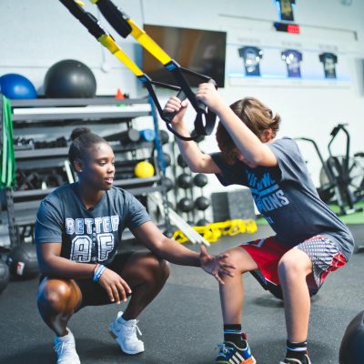 A teenager is working out using resistance bands while being guided by his coach in a gym.